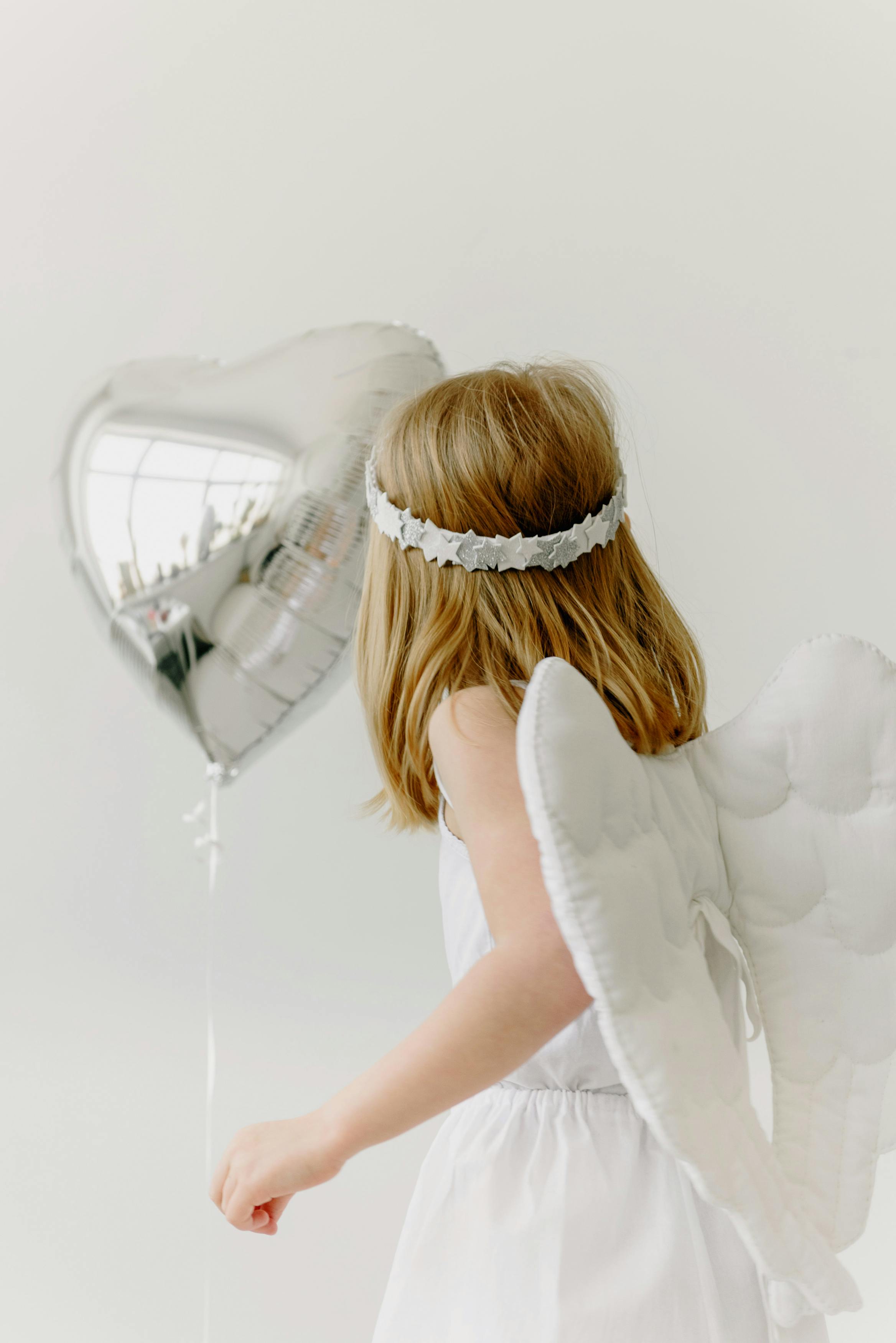 A child in an angel costume holding a heart-shaped silver balloon in a studio setting.