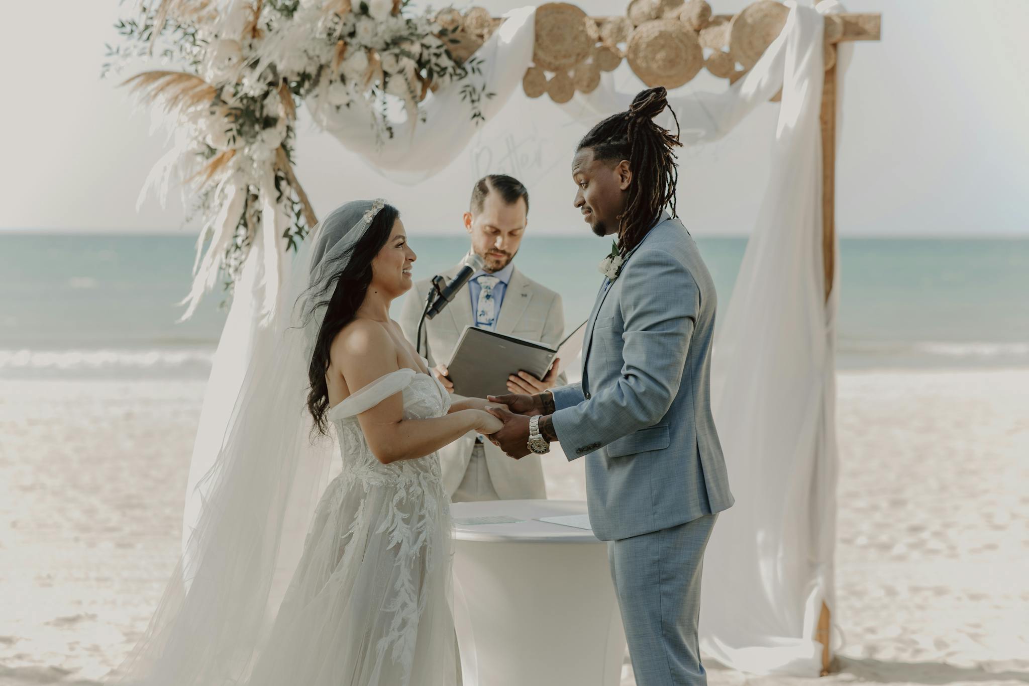 A romantic beach wedding ceremony with a bride and groom holding hands, guided by an officiant.
